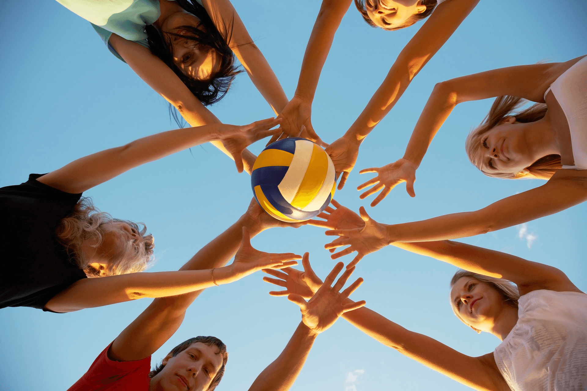 Group of people reaching together for a volleyball against a blue sky