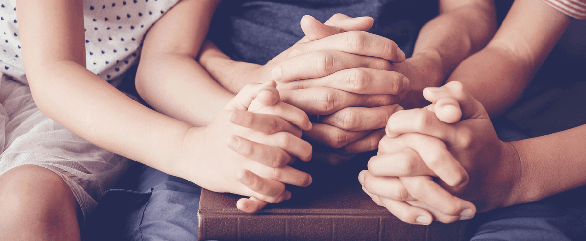 Family hands clasped in prayer together over a Bible